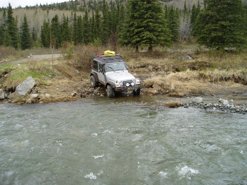 Silver Jeep Wrangler driving through a shallow stream with trees and a hill in the background
