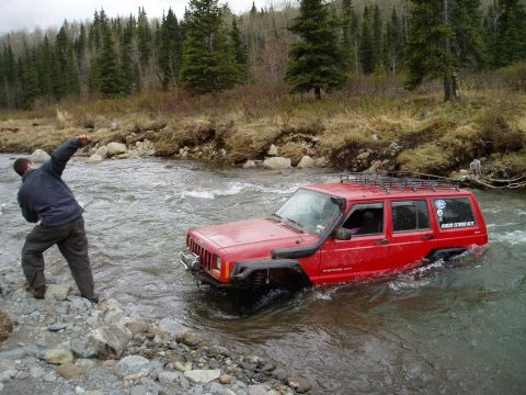 2000 Ford E350 - Jeep Cherokee XJ navigates a river crossing with a person watching from the bank.