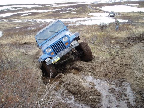2000 Ford E350 - A blue Jeep Wrangler navigates a muddy off-road course with other vehicles in the background.