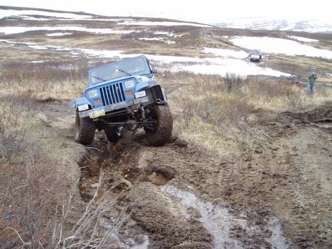 2000 Ford E350 - A blue Jeep Wrangler tackles a challenging muddy terrain, with a second vehicle visible further up the trail.