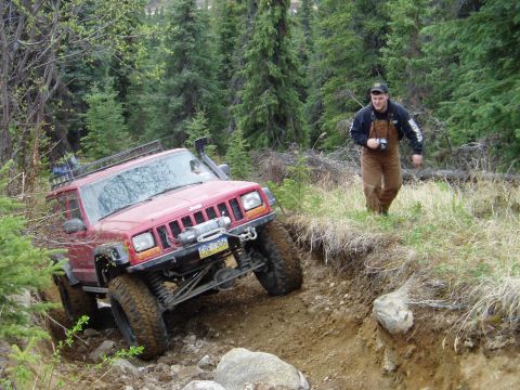2000 Ford E350 - A red Jeep Cherokee navigates a challenging off-road trail with a man walking nearby.