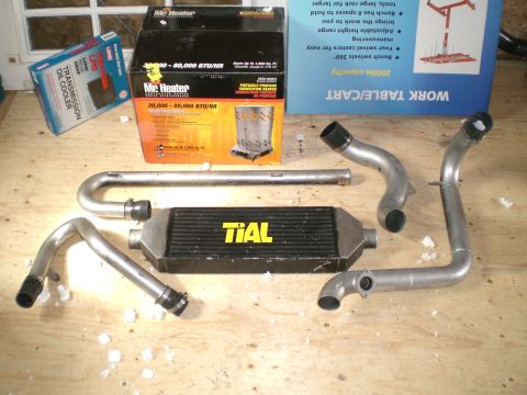 1995 Ford Probe - A black bar-and-plate TiAL front-mount intercooler with yellow logo laid out on a plywood bench alongside four mandrel-bent aluminum charge…