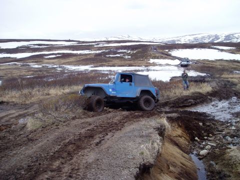 2000 Ford E350 - A blue Jeep Wrangler tackles a challenging muddy terrain, with other vehicles visible in the distance under a cloudy sky.