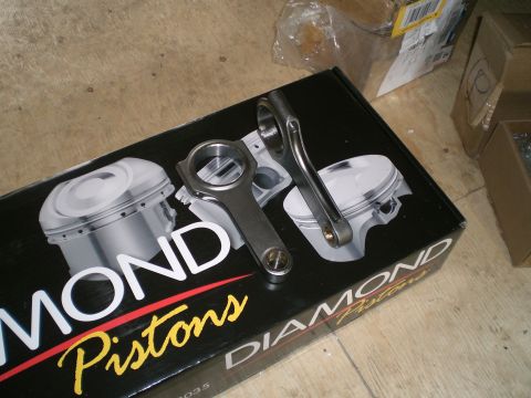1995 Ford Probe - Two forged H-beam connecting rods with bronze wrist-pin bushings resting on top of a black Diamond Pistons box on a plywood floor, with shi…