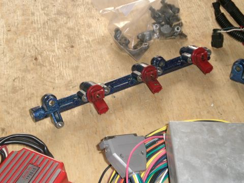 1995 Ford Probe - Blue metal-flake powder-coated fuel rail with four red-bodied fuel injectors clipped in place, laid on a plywood workbench beside a bag of…