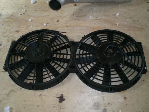 1995 Ford Probe - Dual black electric radiator cooling fans joined side by side in a plastic shroud, laid face-up on a plywood bench with their blue and blac…