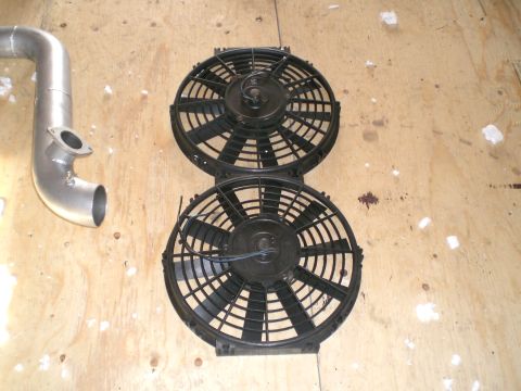 1995 Ford Probe - A pair of black S-blade electric radiator cooling fans with plastic shrouds and pigtail wiring lying side by side on a plywood floor. A bar…