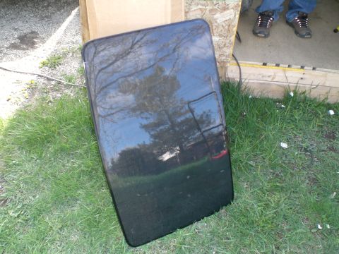 1995 Ford Probe - A removed tinted glass sunroof panel with a black frame and dotted ceramic frit border, propped upright in the grass against a cardboard sh…