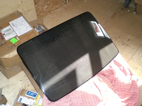 1995 Ford Probe - A removed tinted glass sunroof or moonroof panel laid face-up on a plywood shop floor, its black perimeter seal and dot-matrix ceramic frit…