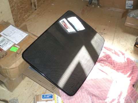 1995 Ford Probe - Glossy black carbon fiber panel with a fine plain-weave pattern and rounded corners, propped at an angle on a plywood floor and reflecting…