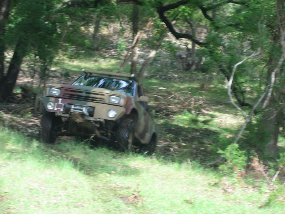 Camo-painted lifted pickup with winch bumper climbing a grassy slope on a wooded trail