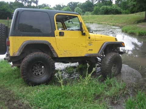 2001 Jeep Wrangler - A yellow Jeep Wrangler navigates through a muddy, watery terrain with lush green grass and trees in the background.