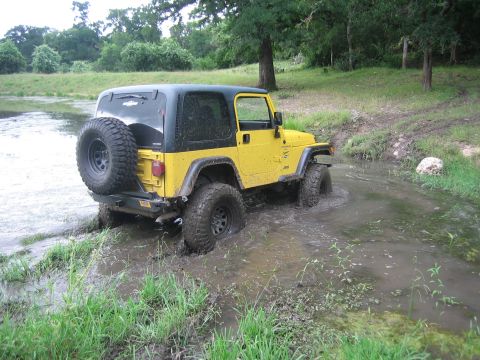 2001 Jeep Wrangler - A yellow 2001 Jeep Wrangler navigates a muddy, water-filled path surrounded by greenery.