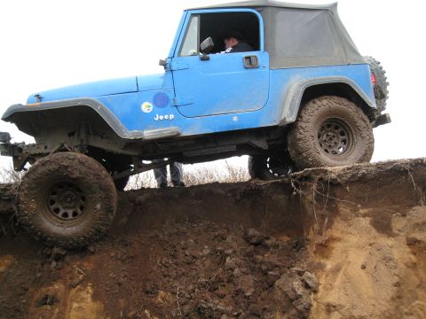 2000 Ford E350 - A blue Jeep Wrangler navigates a challenging dirt obstacle, with its front tires on the edge of a drop-off.