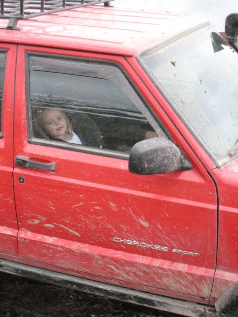 2000 Ford E350 - A child smiles from the window of a mud-splattered red Jeep Cherokee Sport.