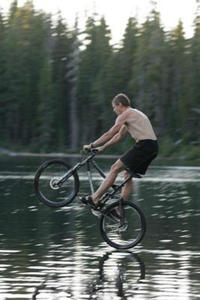 1986 Ford Bronco - A man rides a bicycle on one wheel across a body of water, with a forest behind him.