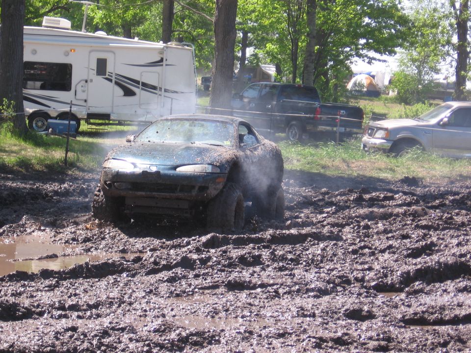 Blue sports car struggling in deep mud with a trailer and trucks in background