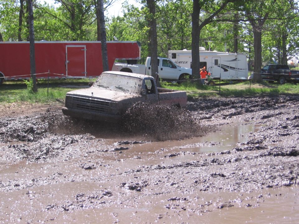 Mud-covered pickup truck splashing through a muddy track with trees and trailers in the background