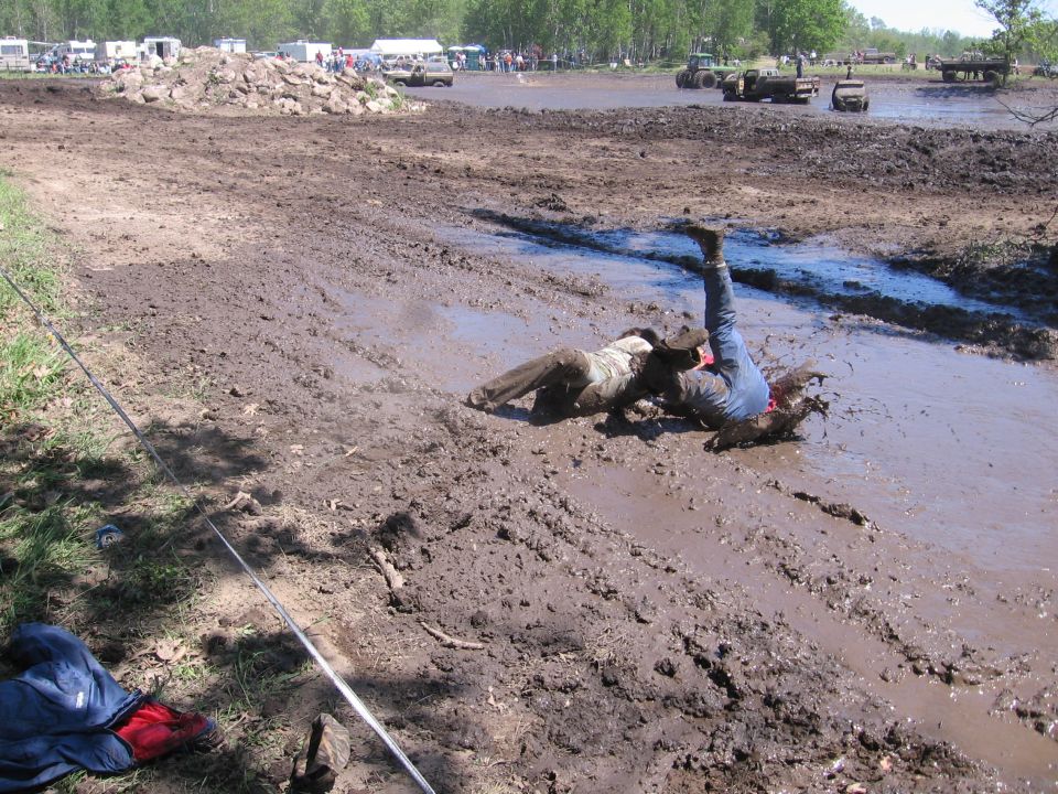 Two people are in a mud pit with vehicles and spectators in the background
