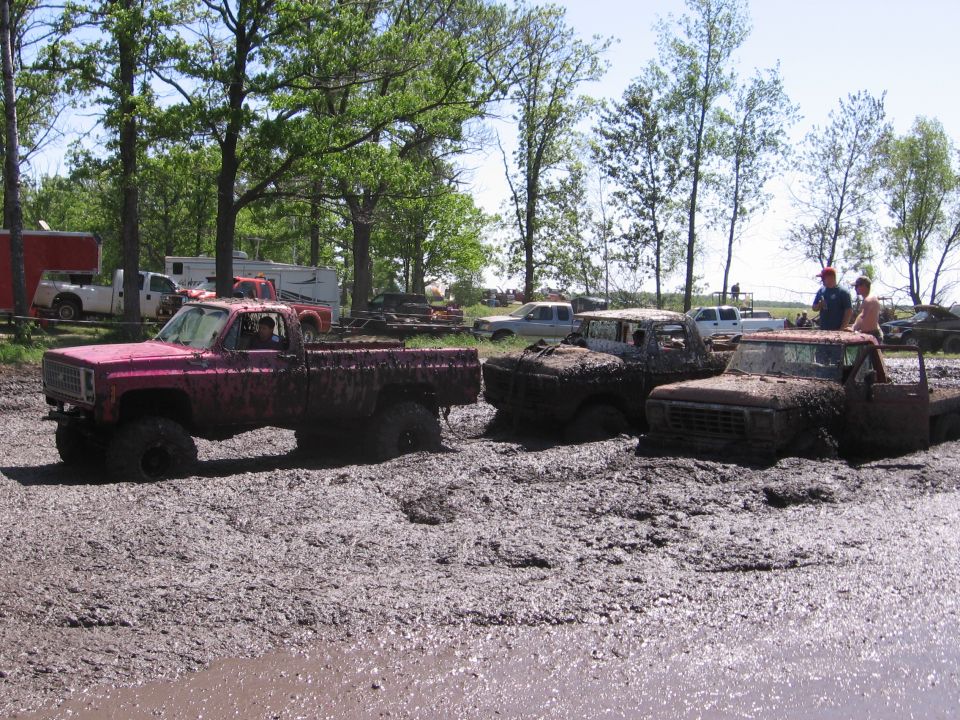 Several trucks stuck in a mud pit with trees and trailers in the background