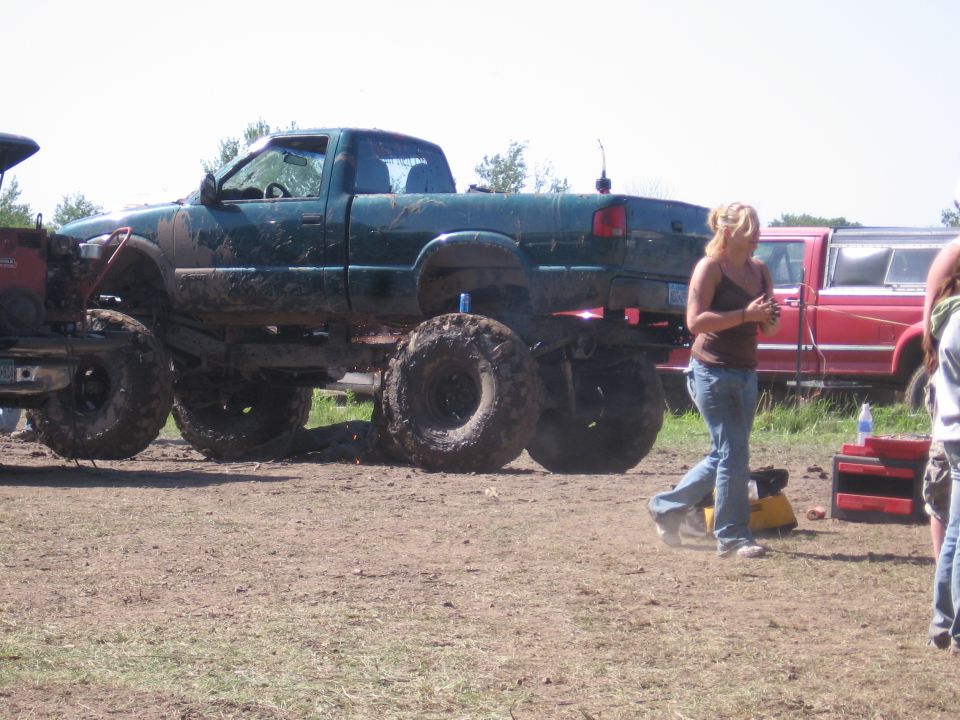 Mud-covered green pickup truck with oversized tires and a woman walking by