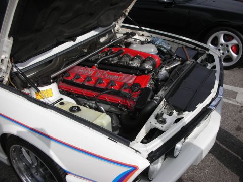 1998 Ford Crown Victoria - Engine bay of a white BMW, displaying its red M Power engine, with a black car visible in the background.