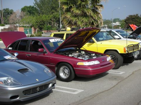 1998 Ford Crown Victoria - A line of cars, including a maroon sedan with its hood up, are parked in a lot on a sunny day.