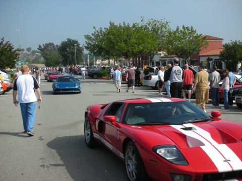 1998 Ford Crown Victoria - A red Ford GT with white racing stripes is parked at a car meet, with other vehicles and people in the background.