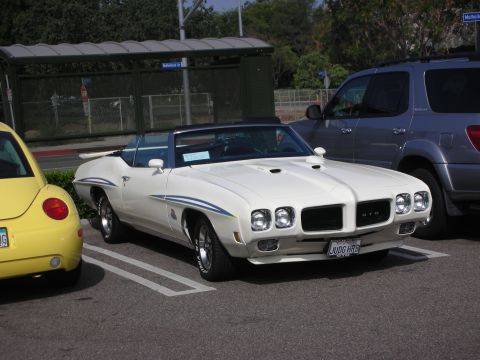 1998 Ford Crown Victoria - A white 1970 Pontiac GTO convertible, distinguished by its blue racing stripes, is parked in a lot alongside a yellow Volkswagen Beetle and…