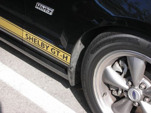 1998 Ford Crown Victoria - A close-up view of the side of a black Shelby GT-H Mustang, showing its distinctive stripe and a Hertz badge.