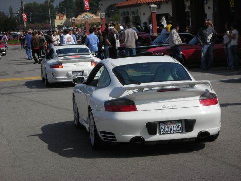 1998 Ford Crown Victoria - A white Porsche 911 is seen from the rear, parked on a street with other vehicles and onlookers.