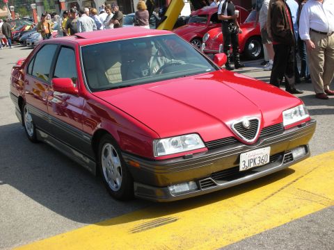 1998 Ford Crown Victoria - A red Alfa Romeo sedan is parked on a street with a yellow line, while onlookers gather.