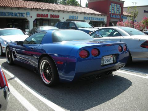 1998 Ford Crown Victoria - A blue Chevrolet Corvette is parked in a lot, with other vehicles and a strip mall in the background.