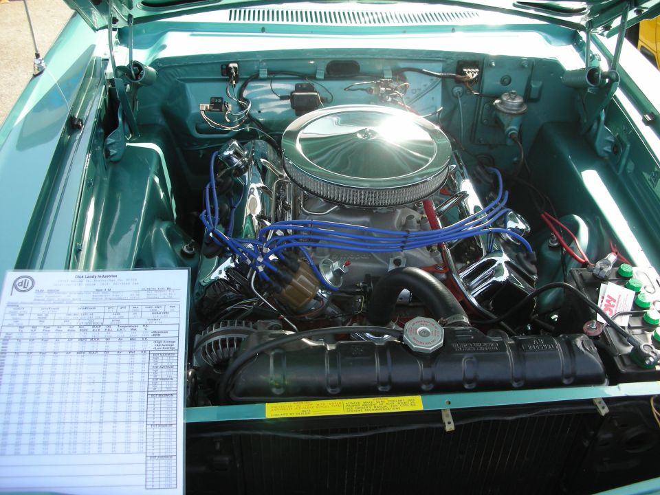 Engine bay of a classic car, showing a V8 engine with chrome air cleaner and blue spark plug wires. A document is on the left