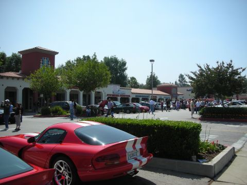 1998 Ford Crown Victoria - A gathering of cars and people in a parking lot outside a strip mall on a sunny day.