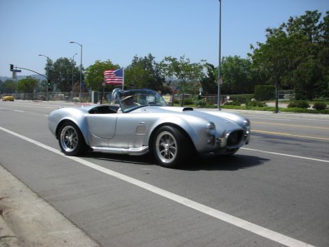 1998 Ford Crown Victoria - Silver AC Cobra replica with an American flag drives down a street.