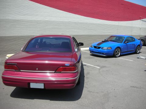 1998 Ford Crown Victoria - A red 1998 Ford Crown Victoria is parked in a lot next to a blue Ford Mustang.