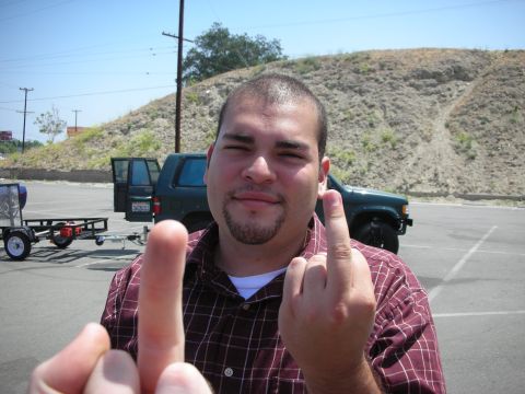 1998 Ford Crown Victoria - A man provocatively gestures with both hands while a dark green SUV and trailer sit in the background.