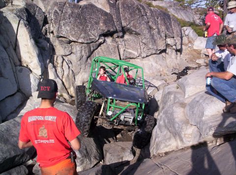 1989 Ford Bronco - A green tube-frame rock buggy with two occupants in red shirts climbs a granite ledge on a rocky off-road trail, running a front-mounted wi…