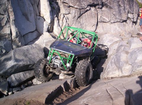 1989 Ford Bronco - A green tube-frame rock buggy with a black hood crawls over large rocks on an off-road trail, carrying two occupants, one wearing a red shi…