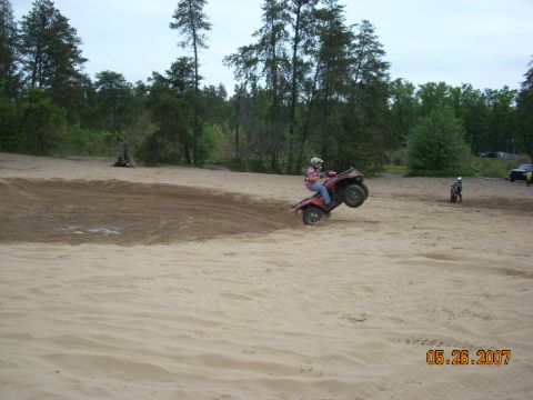 2000 Chevrolet Tahoe - An ATV rider performs a wheelie on a sandy track with trees and another rider in the distance.