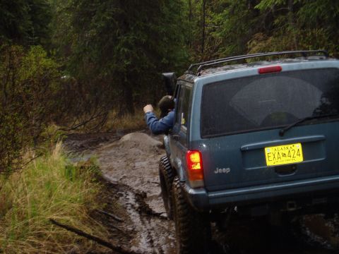 2000 Ford E350 - A teal Jeep Cherokee navigates a muddy off-road trail, with a person visible in the driver's seat.