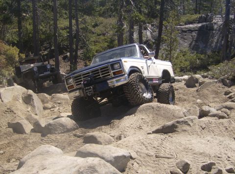 1986 Ford Bronco - Two off-road vehicles navigate a rocky trail in a wooded area.