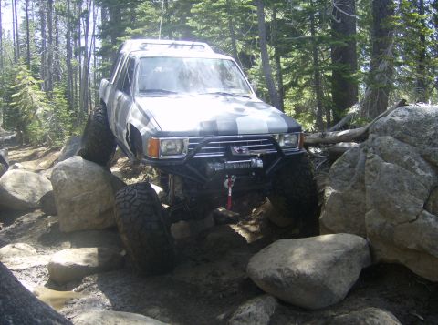 1986 Ford Bronco - This modified SUV is shown off-roading through a challenging rocky path in a wooded area.