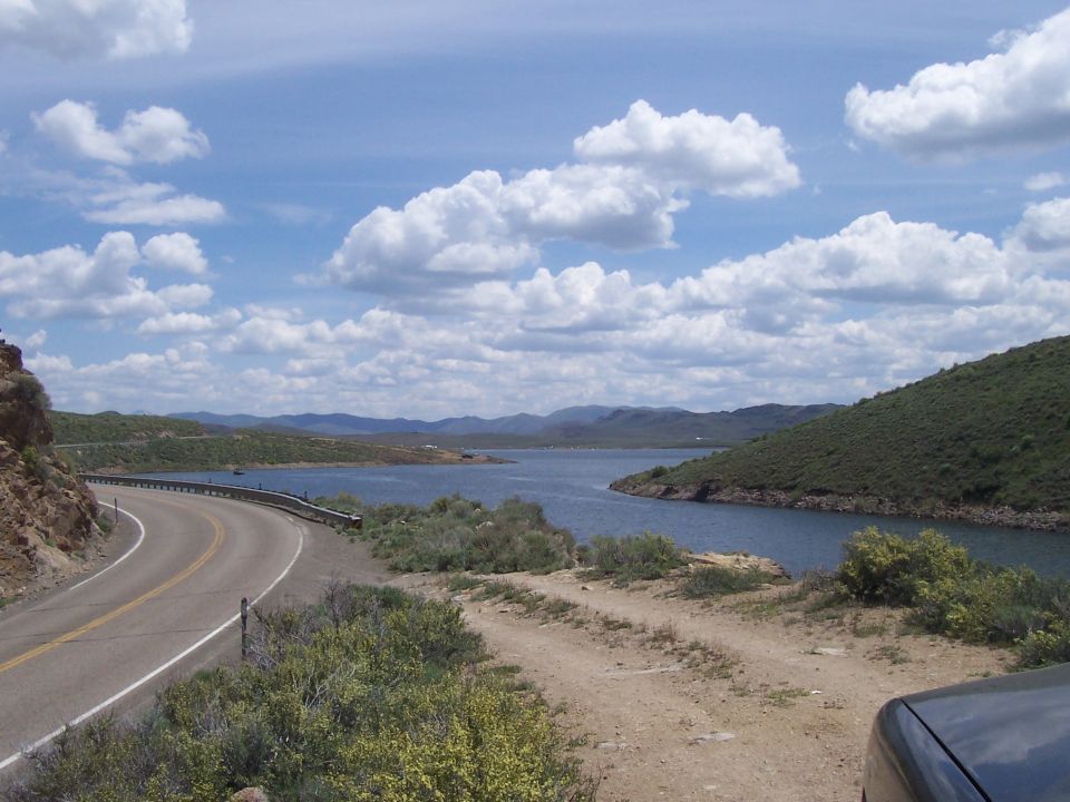 Road curves by a lake with green hills under a cloudy sky. Part of a dark vehicle is in the foreground