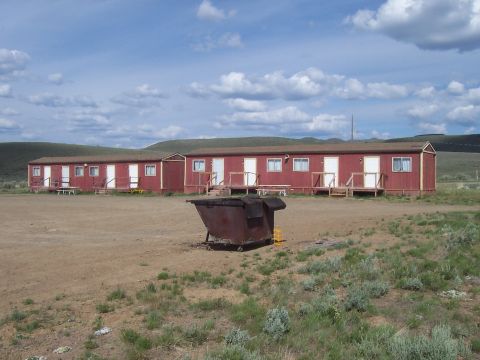 1994 Ford F-150 - A large, rusty dumpster sits in a dirt lot before a long, red building with multiple doors.