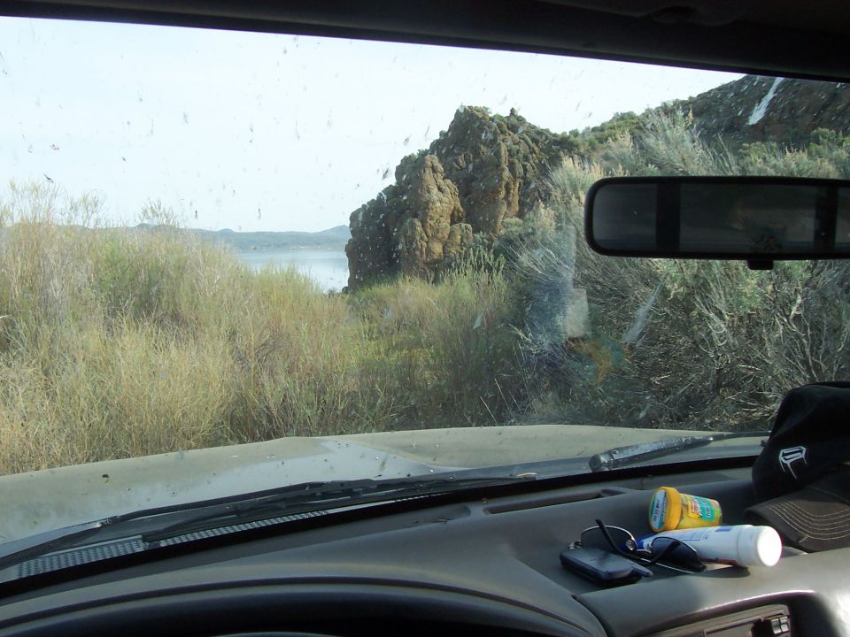 View from inside a vehicle looking out at a lake and rocky hills through a dirty windshield. Sunglasses, keys, and a hat are on the dashboard