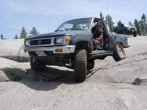 1985 Toyota Hilux - A dark green Toyota pickup truck is shown off-roading on a rocky terrain with another truck in the background.