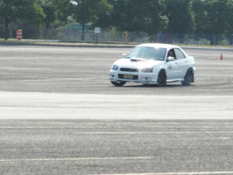 2001 Mercury Grand Marquis - A white Subaru Impreza WRX is captured mid-drift on a paved surface, with trees and signs in the background.