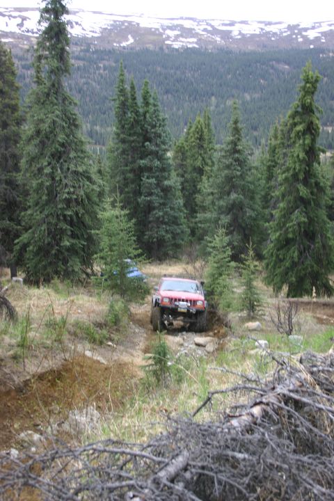 2003 Jeep TJ - A red Jeep Wrangler navigates a rocky trail through a forest with snow-capped mountains in the background.